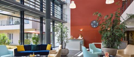 A modern lobby with a reception desk, seating area, large windows, and indoor plants.
