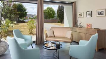 Un salon moderne avec un canapé beige, deux fauteuils bleu clair, une petite table ronde sur laquelle se trouvent une théière et une assiette de pâtisseries, et un meuble en bois orné de cadres avec des photos.