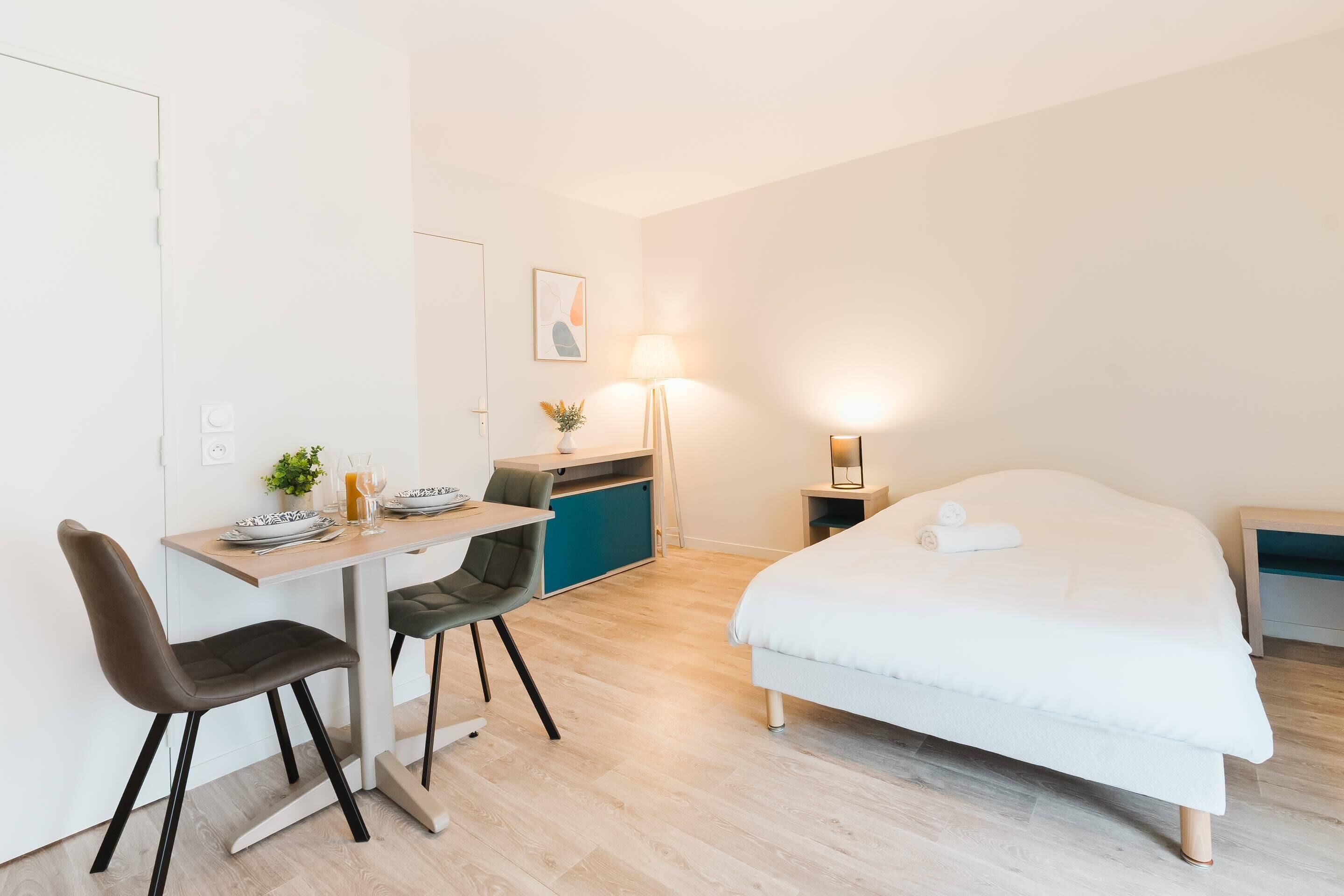 A modern hotel room with a bed, a desk with two chairs, a small kitchenette, and a bedside table with a lamp.
