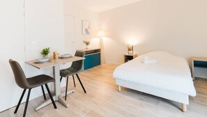 Una habitación de hotel moderna con una cama, un escritorio con dos sillas, una pequeña cocina y una mesita de noche con una lámpara.