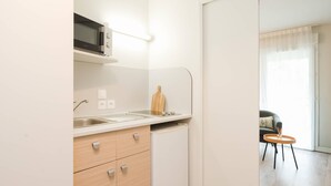 Une cuisine compacte équipée d’un four à micro-ondes, d’un évier et d’un petit réfrigérateur. On y trouve une planche à découper en bois sur le plan de travail et une chaise dans l’espace de vie adjacent.
