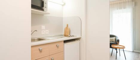 A compact kitchen with a microwave, sink, and a small refrigerator. There is a wooden cutting board on the counter and a chair in the adjacent living area.