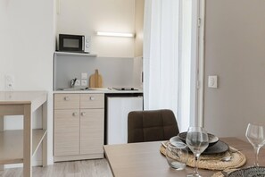 Een compacte keuken met een magnetron, een kleine koelkast en een houten snijplank. Er is een eettafel met een paar borden en glazen.