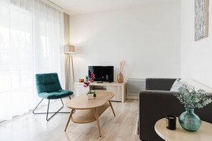 Een moderne woonkamer met een blauwgroene fauteuil, een houten salontafel, een flatscreen televisie, een zwarte bank en een vaas met bloemen.