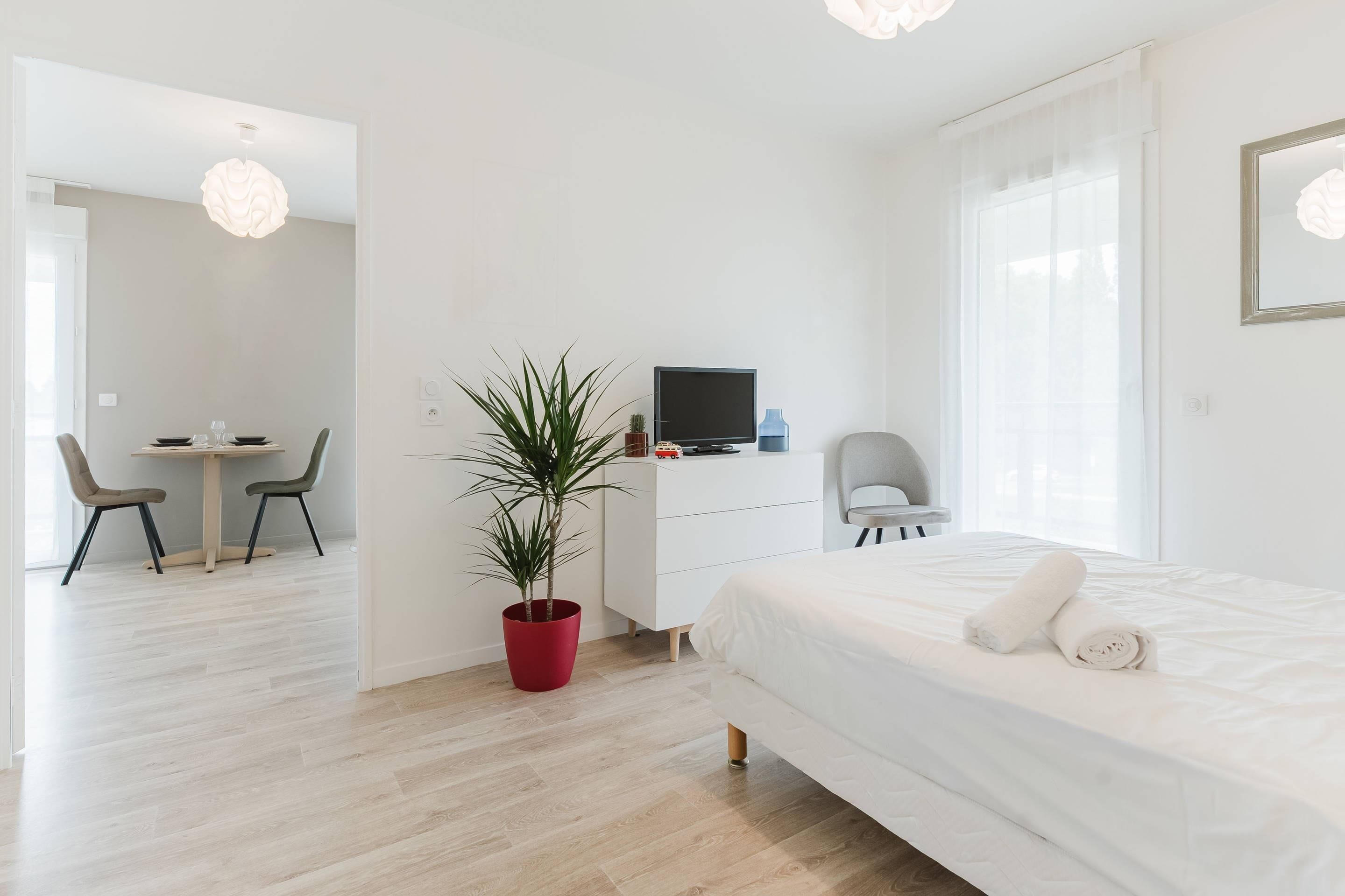 A modern hotel room with a bed, a desk with a computer, a chair, and a potted plant.