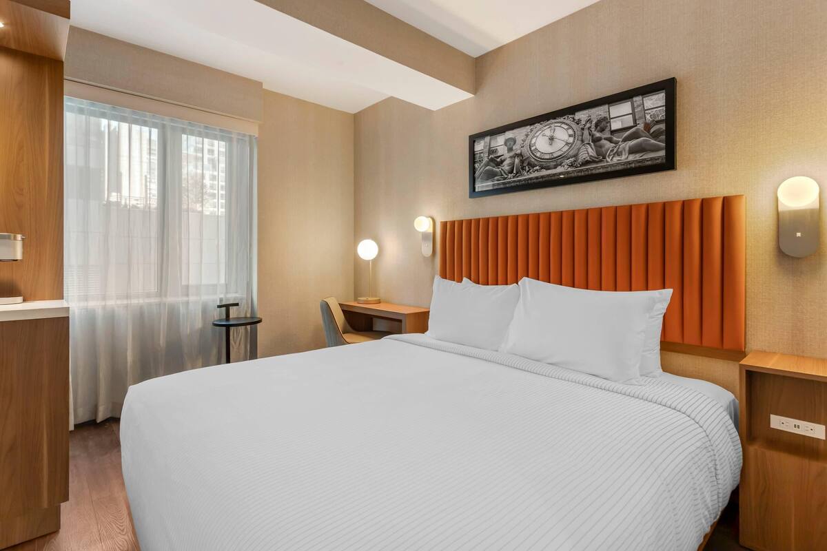 Habitación de hotel con una cama grande, un escritorio, una silla, una ventana con cortinas, una obra de arte colgada en la pared y una lámpara también en la pared.