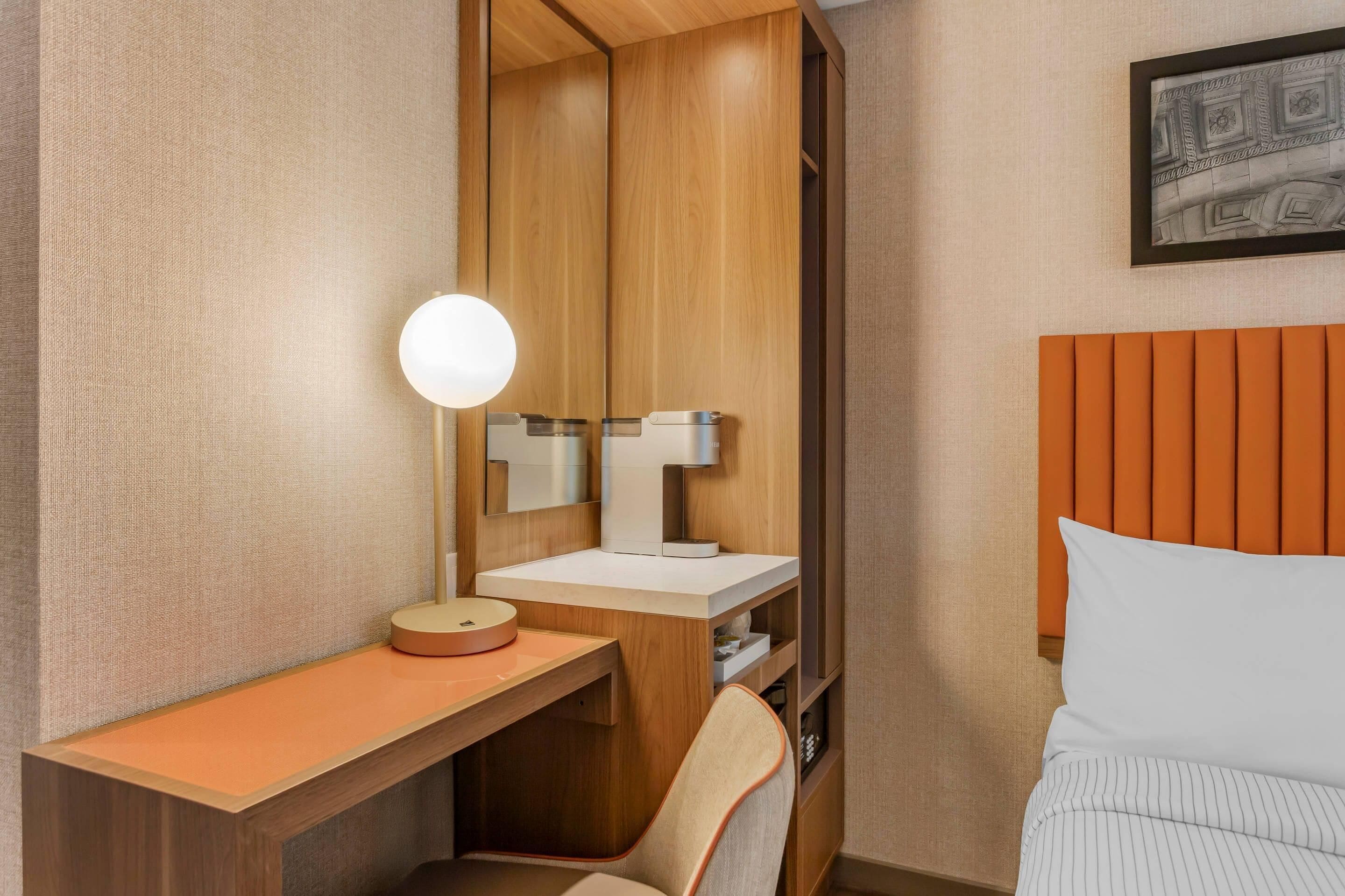 Una habitación de hotel con un escritorio de madera, un espejo redondo, una mesita de noche blanca, una silla y una cama con cabecera naranja.