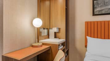 Una habitación de hotel con un escritorio de madera, un espejo redondo, una mesita de noche blanca, una silla y una cama con cabecera naranja.