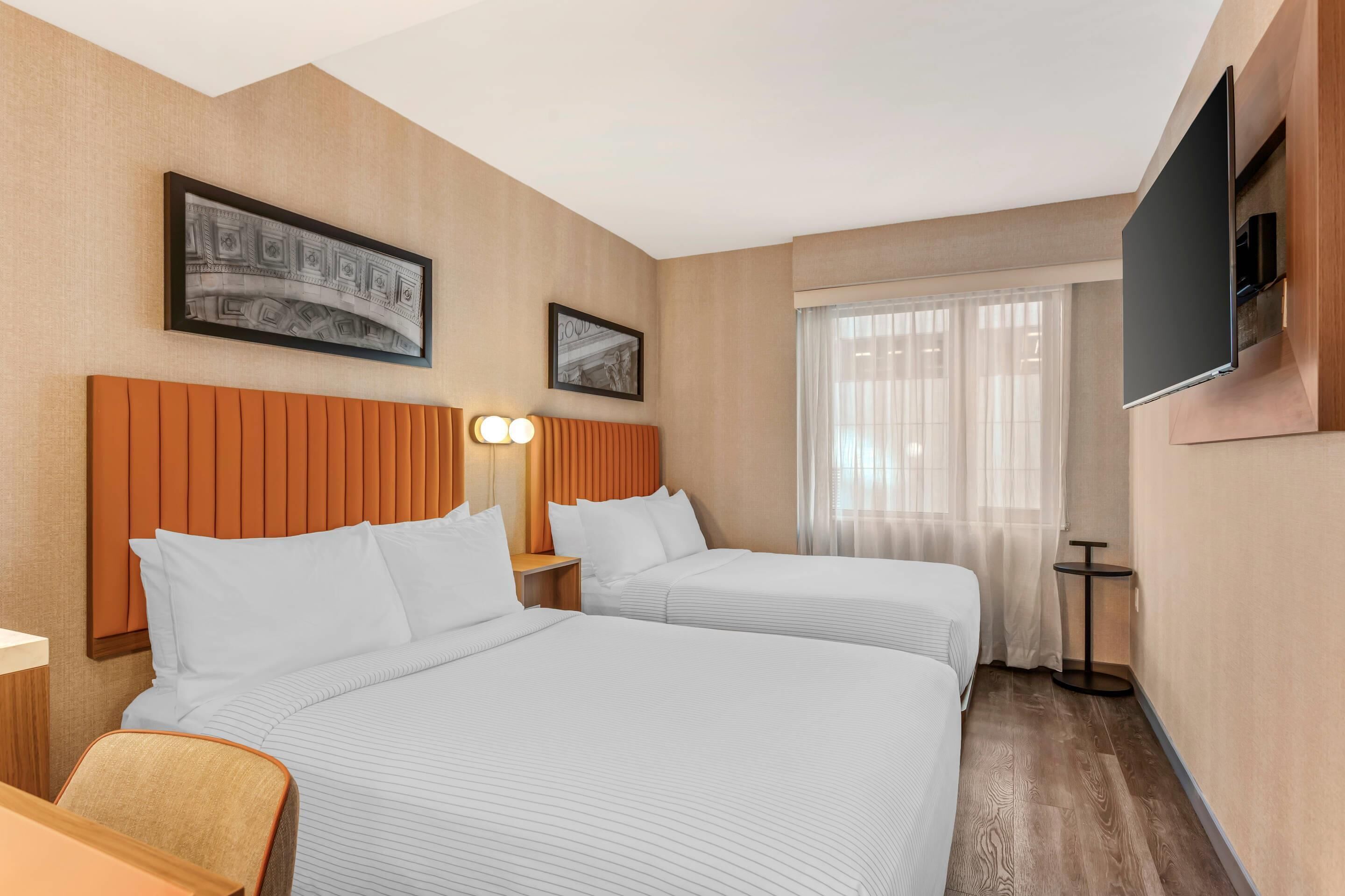 Habitación de hotel con dos camas, un televisor montado en la pared y una ventana con cortinas translúcidas.