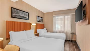 Habitación de hotel con dos camas, un televisor montado en la pared y una ventana con cortinas translúcidas.