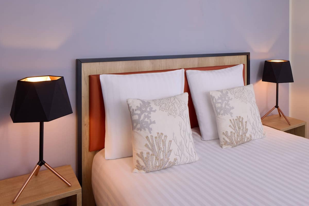 Una cama bien tendida con ropa blanca y dos almohadones decorativos, con dos lámparas negras a cada lado sobre mesitas de noche de madera.