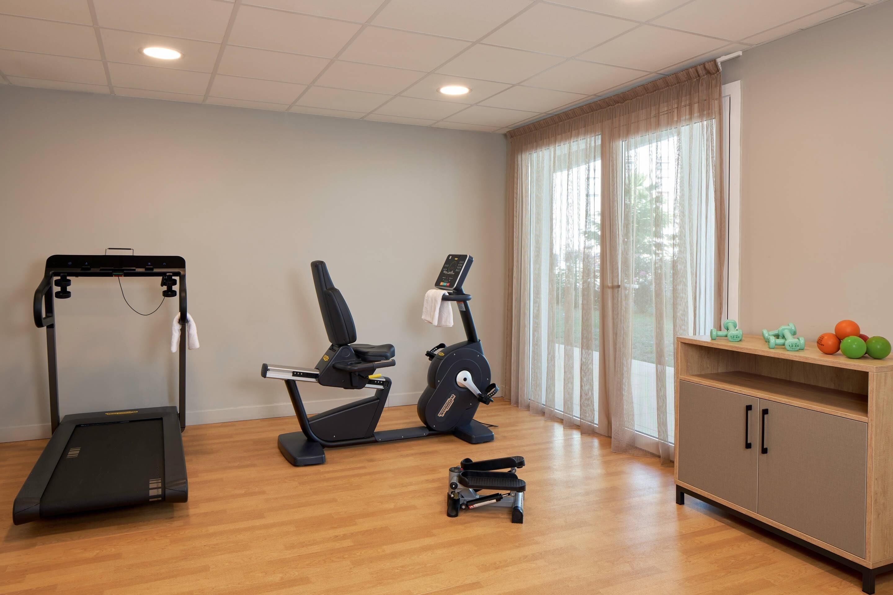 Un gimnasio con cinta de correr, elíptica y bicicleta estática, un armario de madera con mancuernas y una ventana con cortinas.