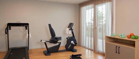 Un gimnasio con cinta de correr, elíptica y bicicleta estática, un armario de madera con mancuernas y una ventana con cortinas.