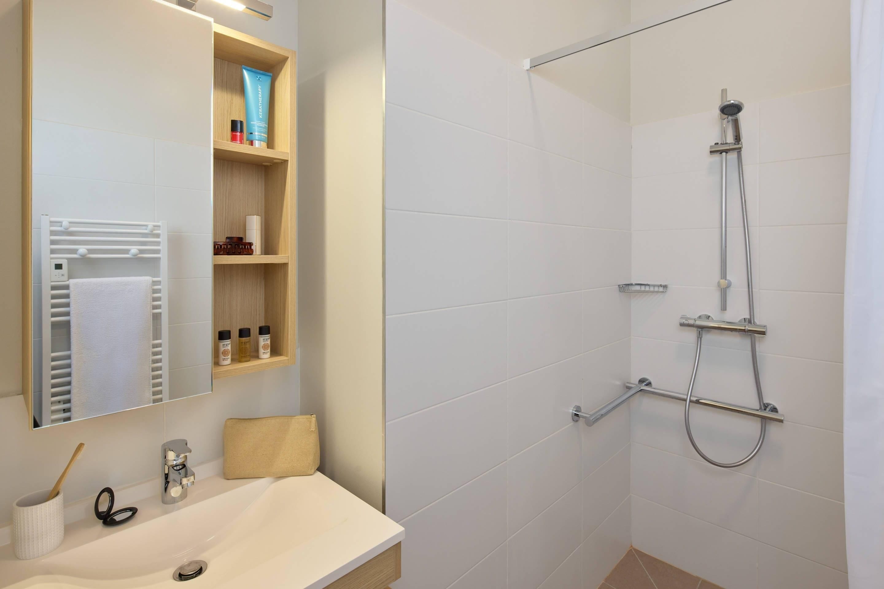 Een moderne badkamer met een witte wasbak, een spiegel, een handdoekrek, een douche met handdouche en een plank met toiletartikelen.
