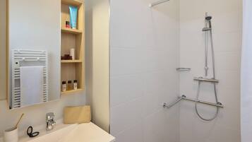 Een moderne badkamer met een witte wasbak, een spiegel, een handdoekrek, een douche met handdouche en een plank met toiletartikelen.
