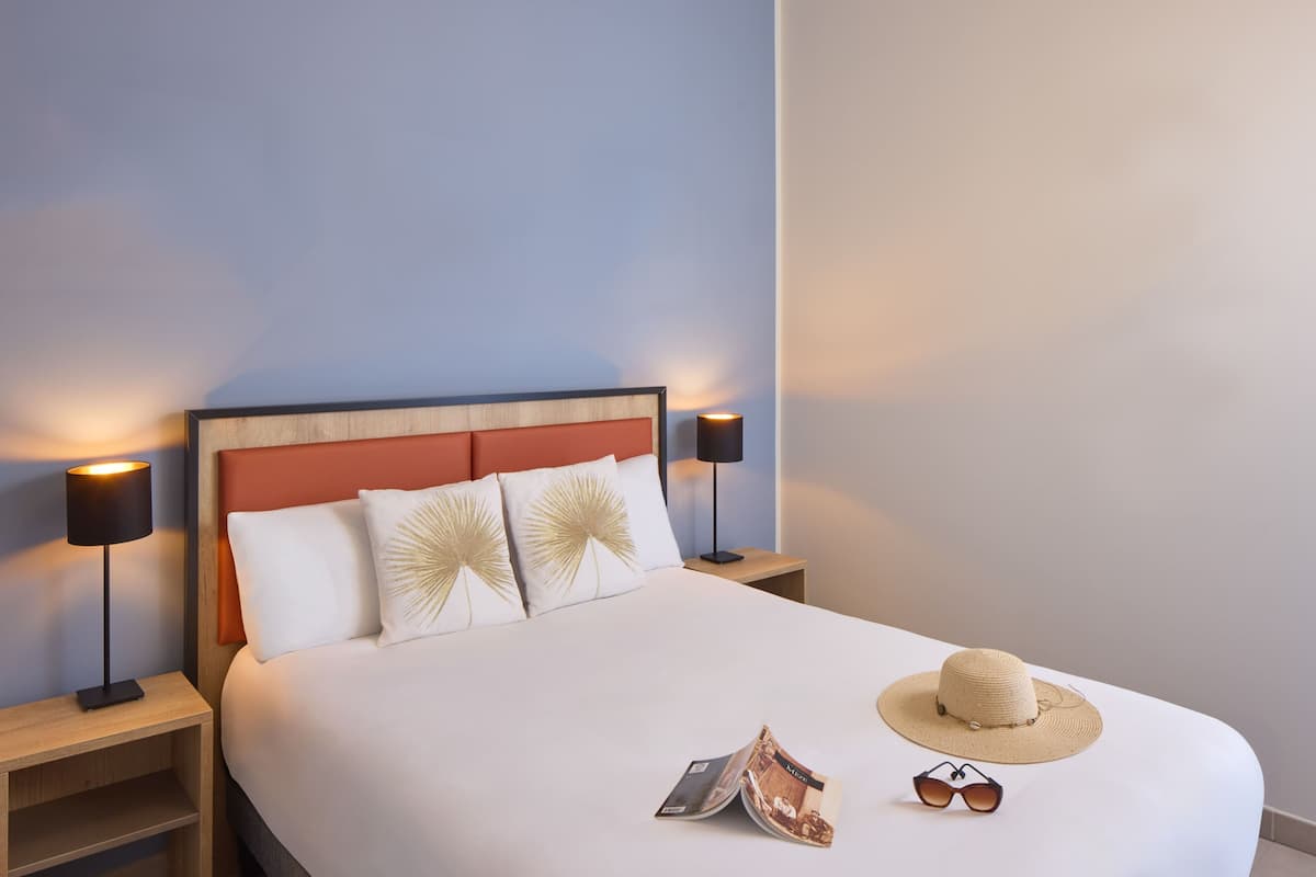 Una cama bien hecha con un cabecero de madera, dos lámparas de noche, un sombrero y gafas de sol sobre la cama.