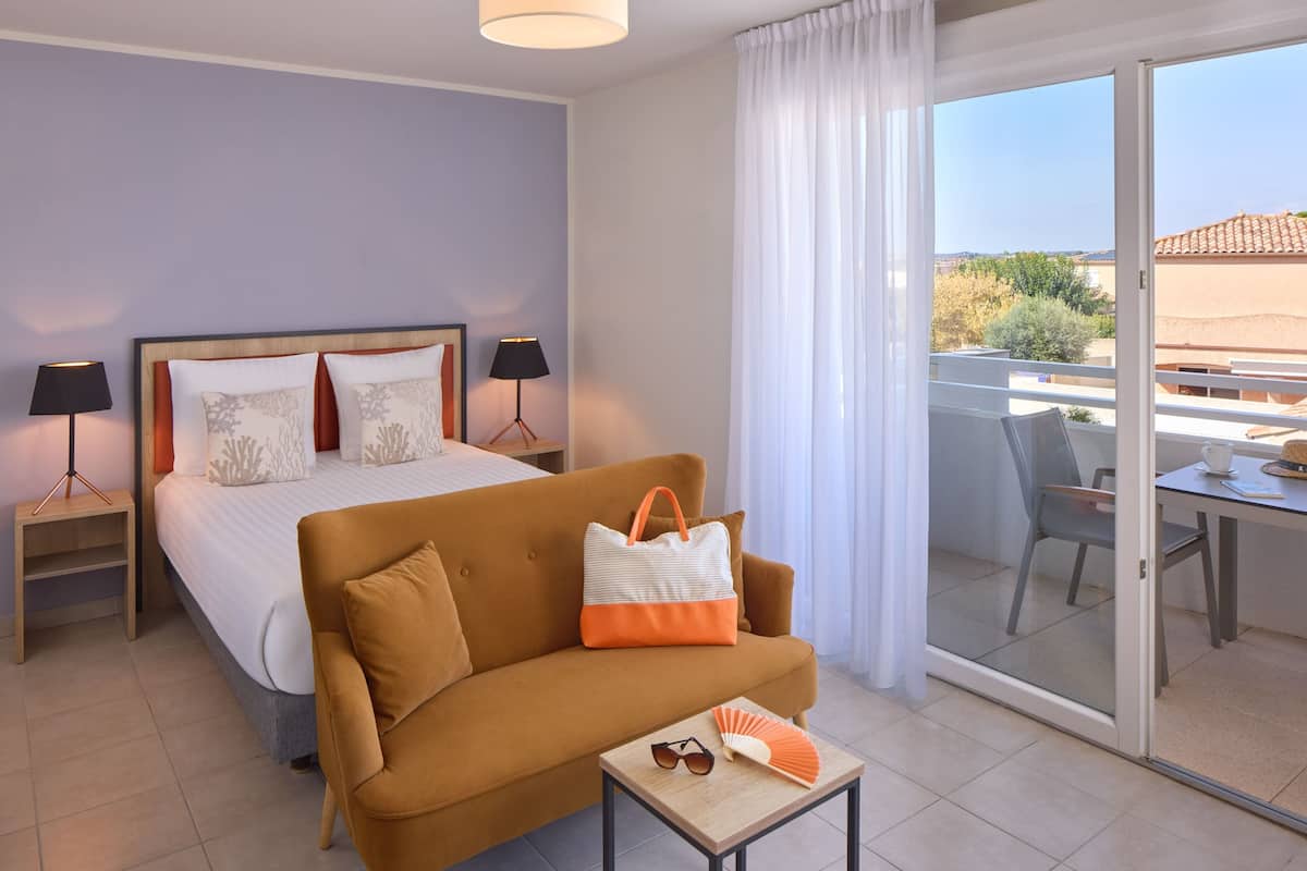 Una habitación de hotel con una cama, un sofá, una mesa con un ventilador y gafas de sol, una mesita de noche con lámparas, y un balcón con una mesa y sillas.