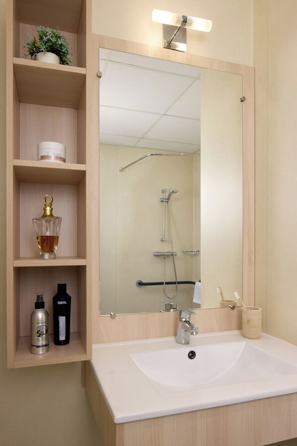 A bathroom with a white sink, a mirror, a shower, and shelves with toiletries. - Quality Aparthotel Perpignan (Perpignan)