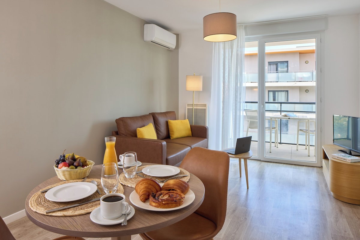 A dining area with a table set for breakfast, including croissants, fruit, and juice. There is a sofa with yellow cushions and a TV on a wooden stand.