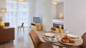 A modern hotel room with a dining area, a sofa, a TV, and a bed.
