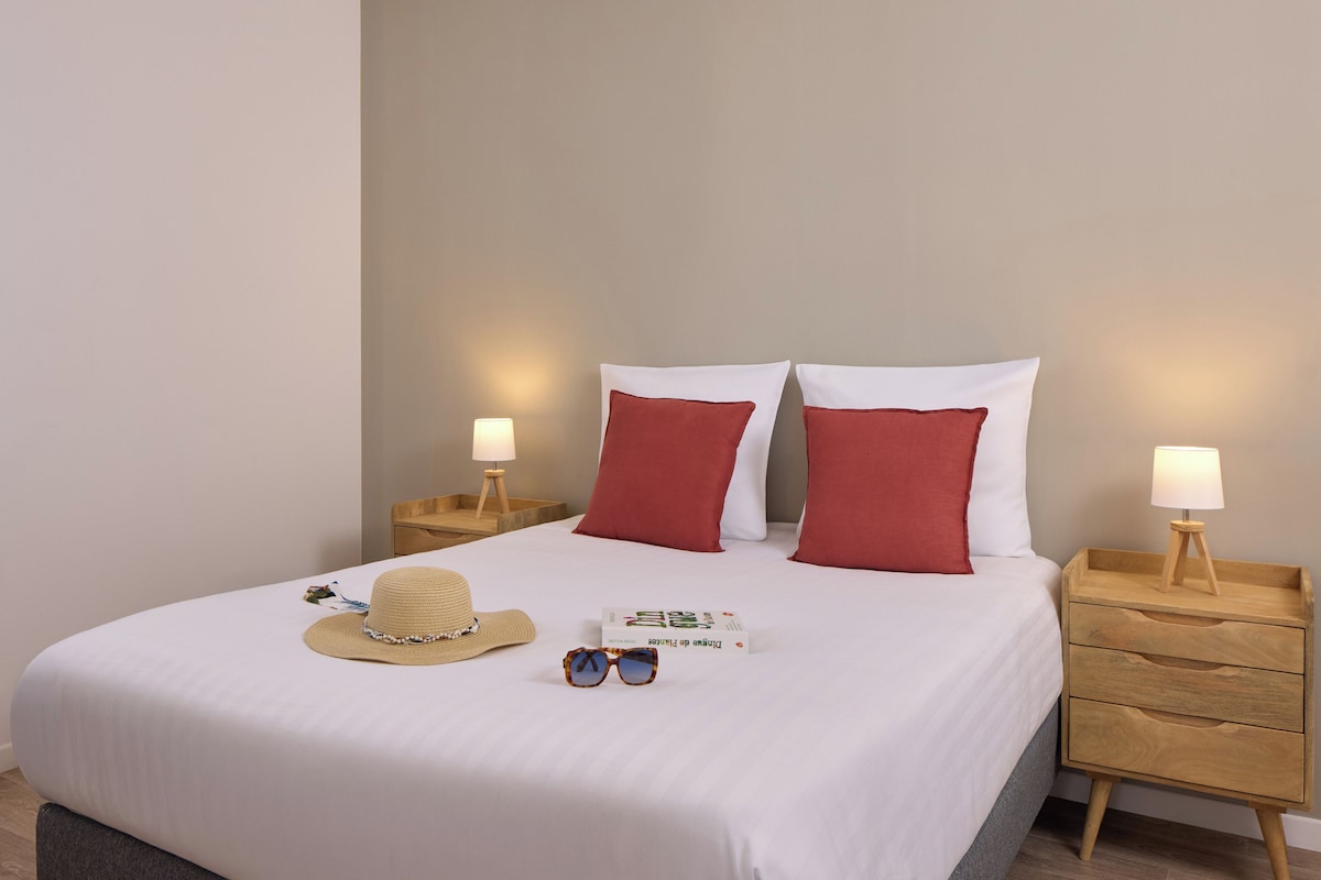 A neatly made bed with white bedding, two red pillows, a beige hat, a pair of sunglasses, and a book on the bed. There are two bedside tables with lamps and a wooden nightstand with drawers.
