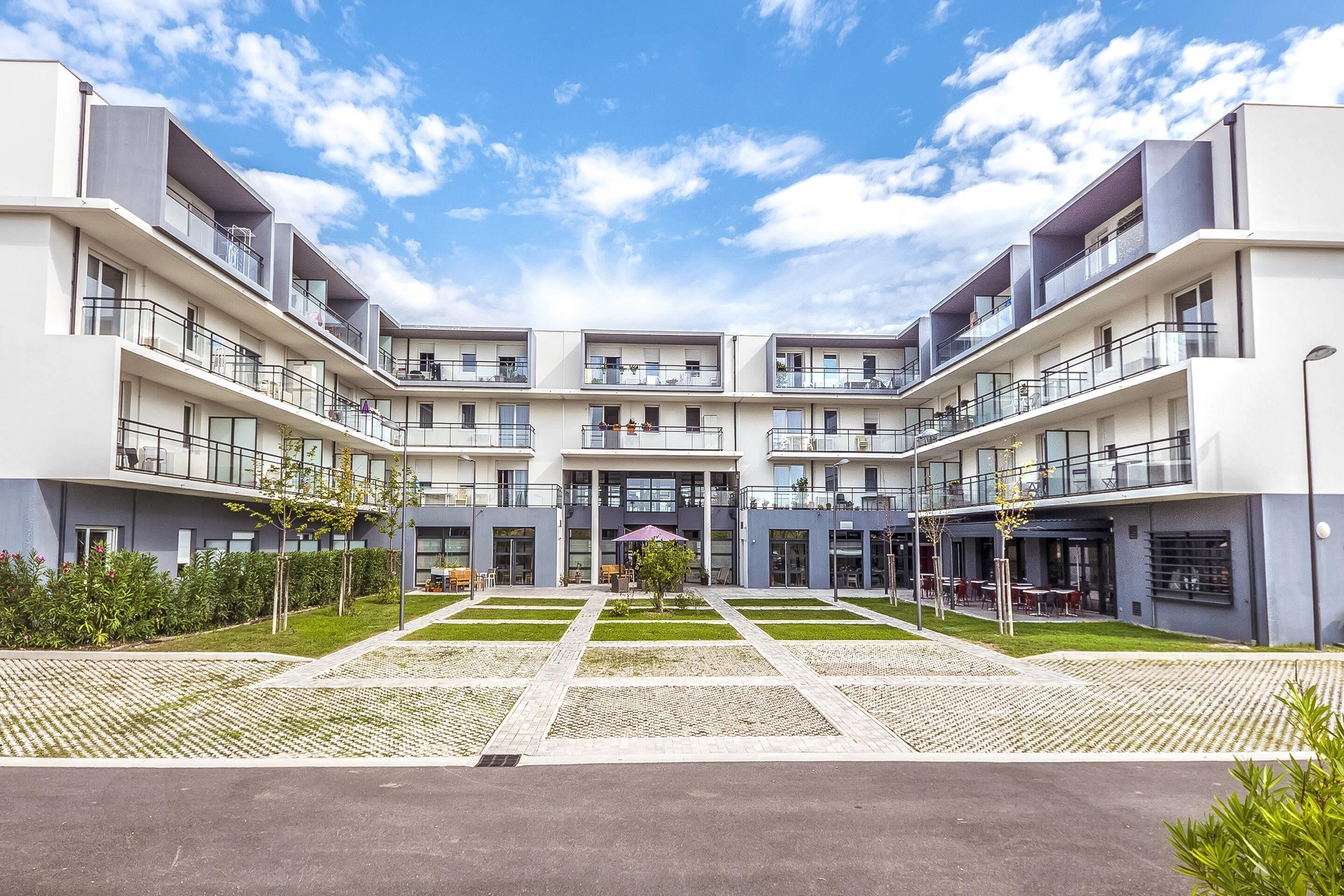 A modern multi-story residential building with balconies, a central courtyard, and a clear sky.