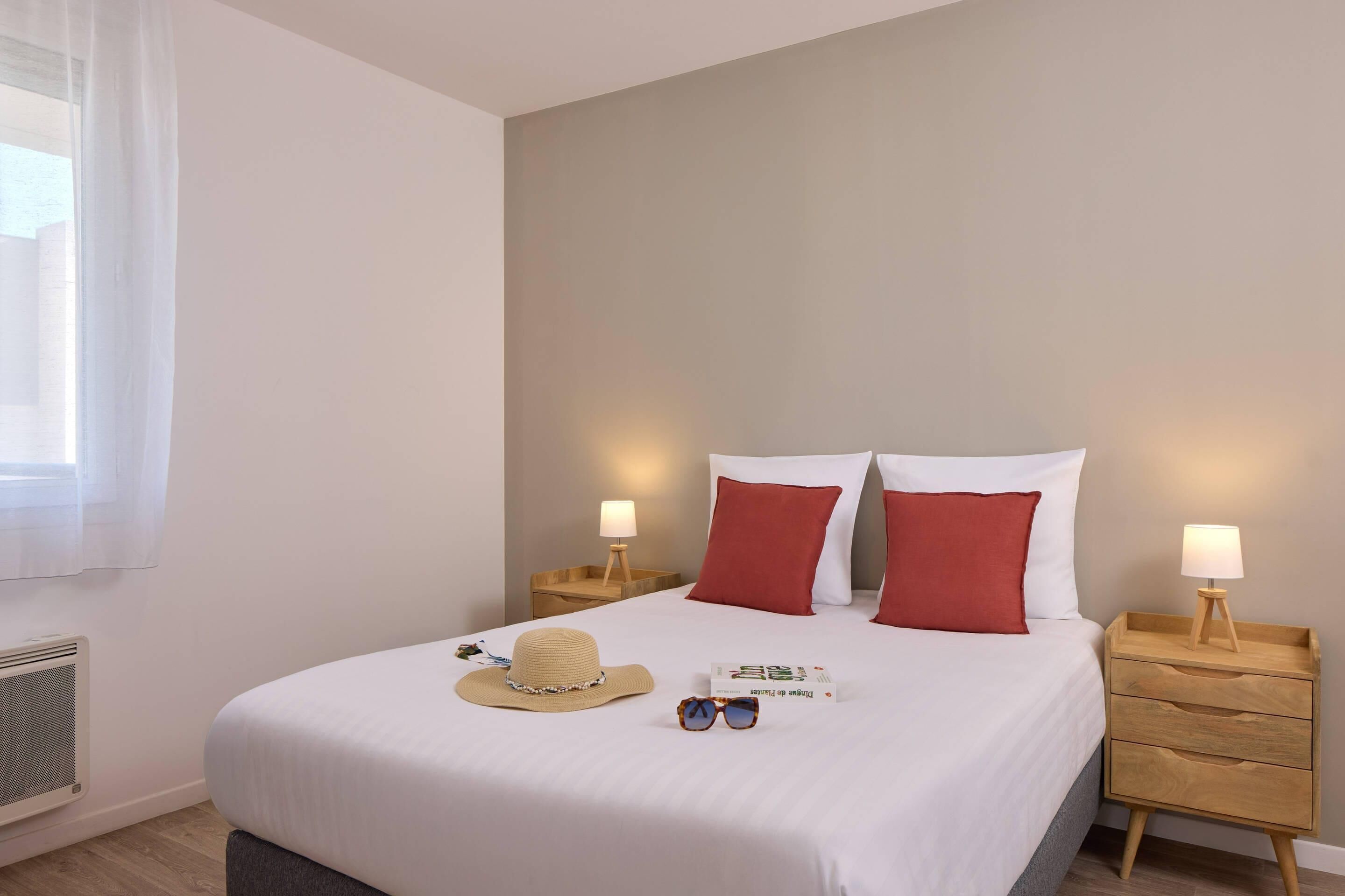 A neatly made bed with white and red pillows, a bedside lamp, a hat, and sunglasses on the bed.