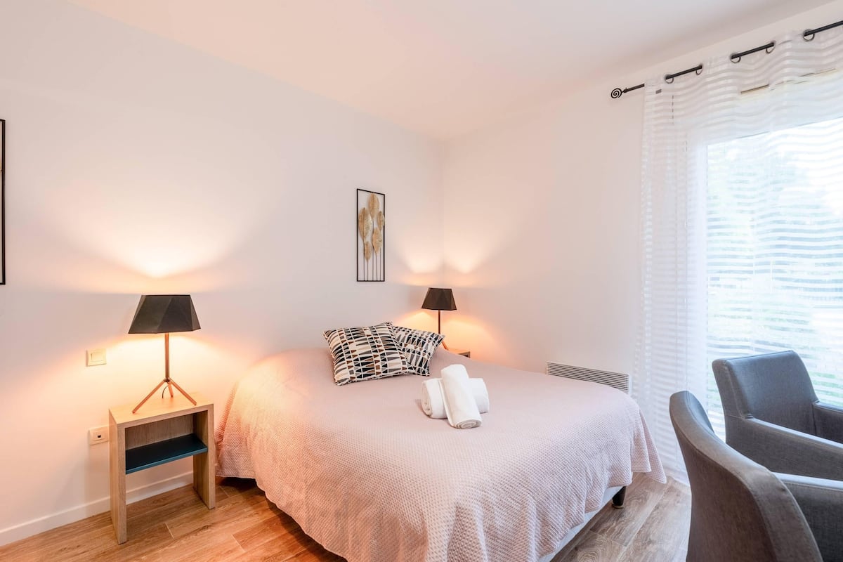 A neatly arranged bedroom with a bed, two bedside lamps, a chair, and a window with curtains.