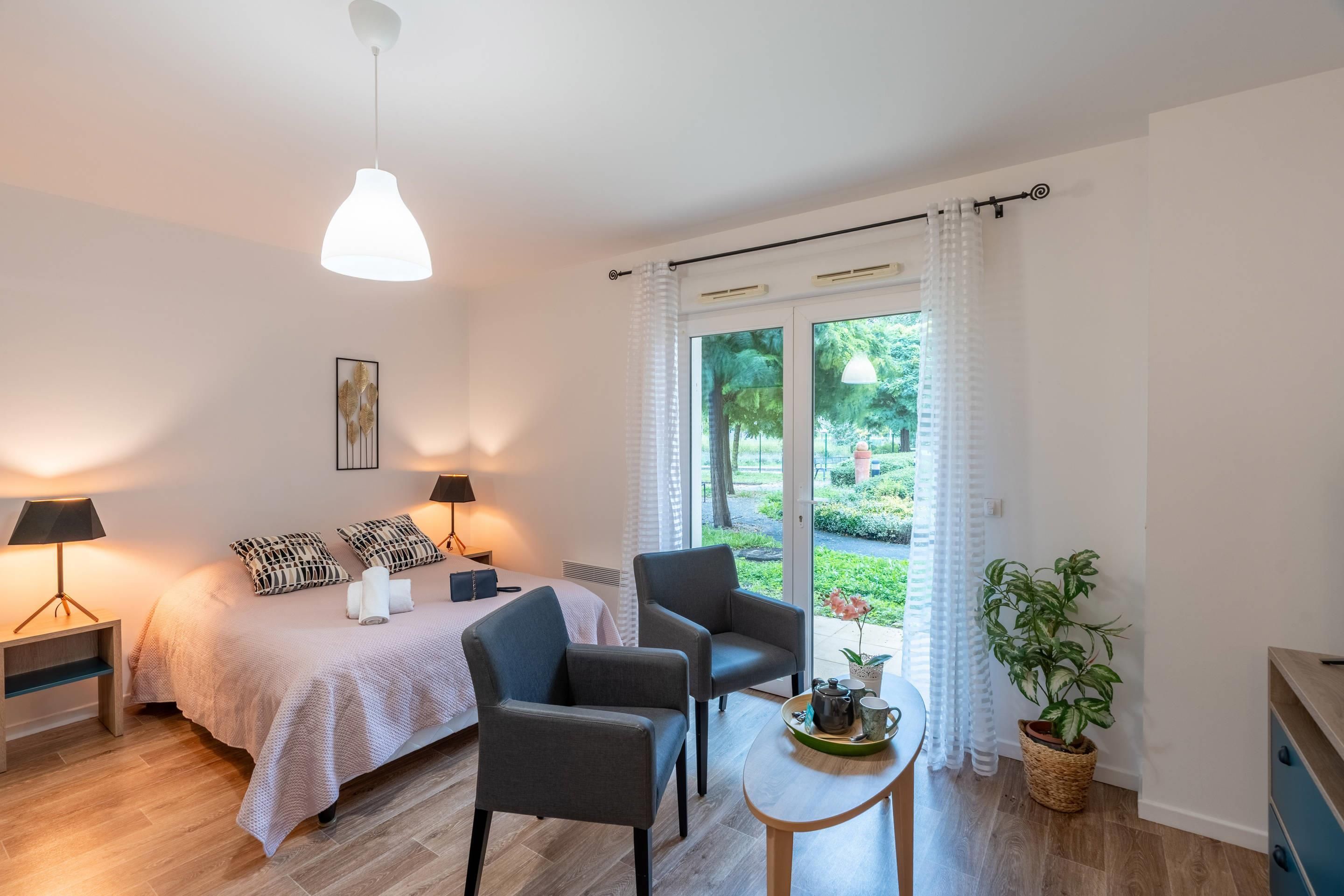 A bedroom with a bed, bedside lamps, a chair, a small table with a tray, and a potted plant.