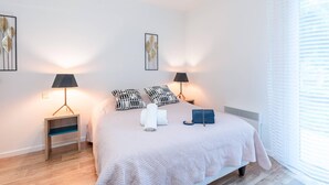 Un dormitorio ordenado con una cama, dos mesitas de noche, una lámpara, un almohadón, una manta y un bolso pequeño.