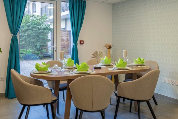 A dining area with a round table set for four, teal curtains, and a view of a building and greenery outside. - Quality Aparthotel Parc Marne la Vallée (Bailly-Romainvilliers)