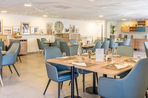 Un espace repas moderne avec des tables préparées pour deux personnes, des chaises bleues et une grande horloge murale.