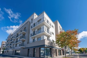 Un immeuble résidentiel à plusieurs étages, doté de balcons, avec un espace commercial au rez-de-chaussée, et un ciel d’un bleu éclatant.