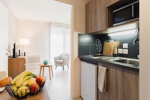 A modern kitchen with a built-in microwave, a sink, and a compact refrigerator. There is a fruit bowl on the counter and a chair in the background. - Quality Aparthotel Dinan (Dinan)