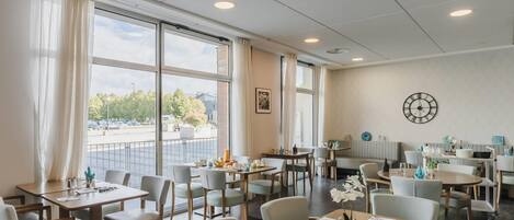 A spacious dining area with multiple tables and chairs, large windows with curtains, a clock on the wall, and a view of the outside.