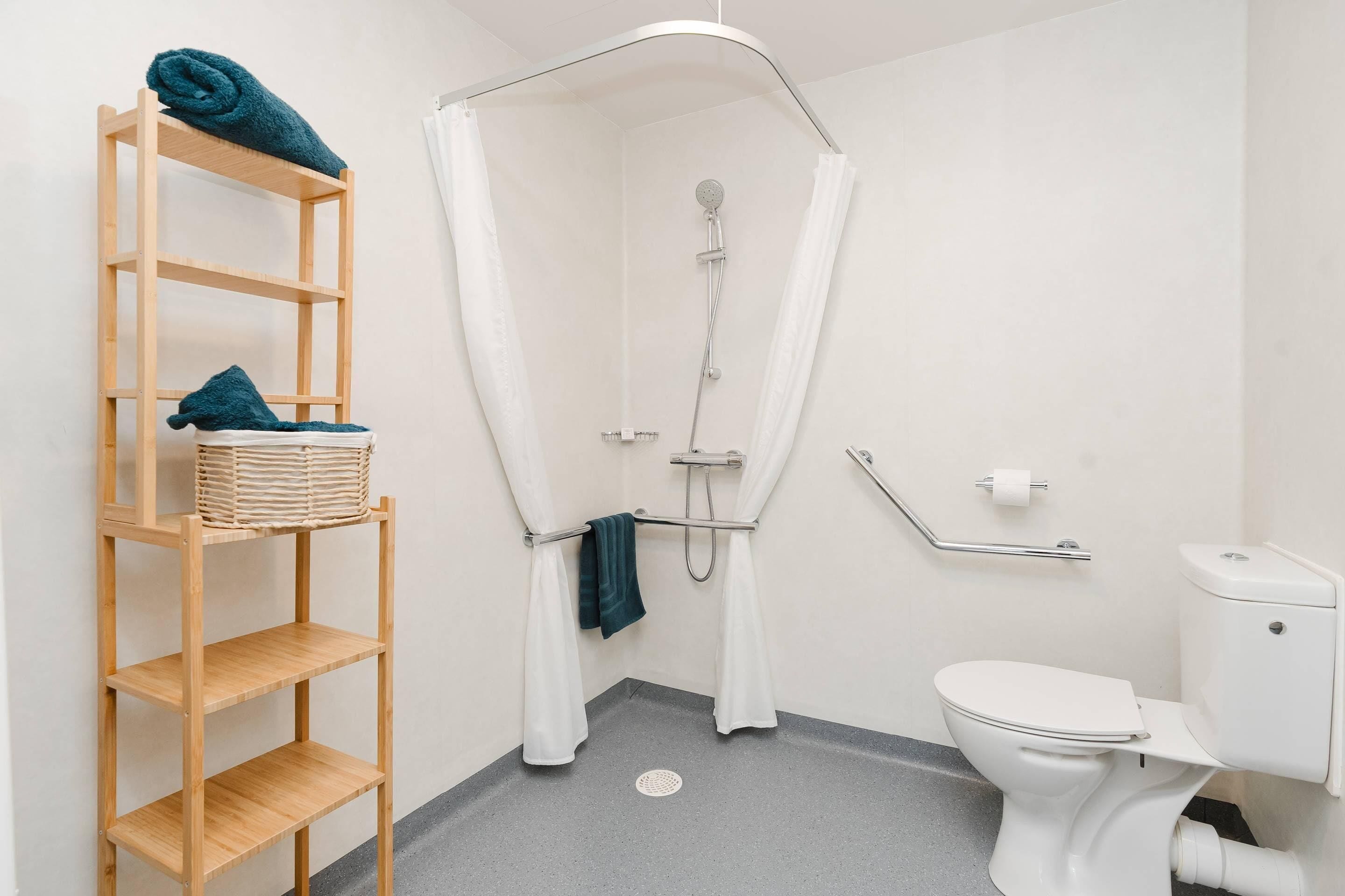 Une salle de bain avec une douche, des toilettes et une étagère en bois où sont rangées les serviettes.