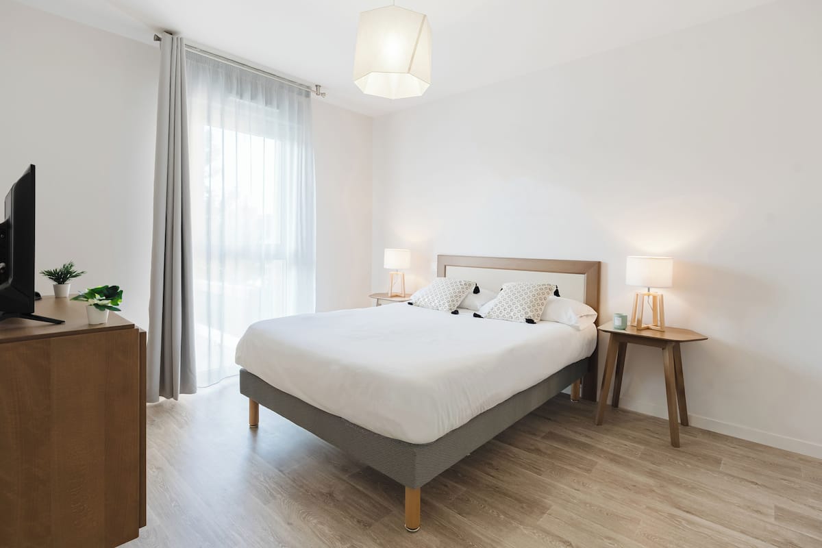 A modern hotel room with a large bed, bedside lamps, a TV on a wooden cabinet, and a window with curtains.