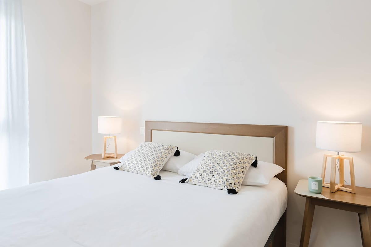 A neatly made bed with white bedding and patterned pillows, flanked by two bedside tables with lamps and a small decorative item.