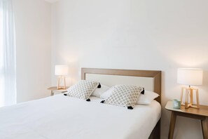 A neatly made bed with white bedding and patterned pillows, flanked by two bedside tables with lamps and a small decorative item. - Quality Aparthotel Dinan (Dinan)