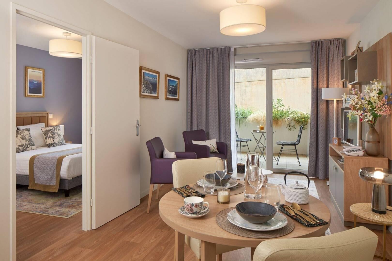 A modern hotel room with a bed, a dining table set for two, a sofa, and a balcony with a table and chairs.