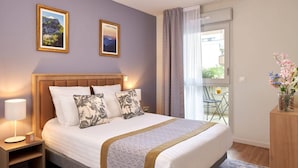 A hotel room with a large bed, two framed pictures on the wall, a bedside table with a lamp, and a view of the outdoors through a window. - Quality Aparthotel Seyssinet (Seyssinet-Pariset)