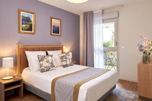 A hotel room with a large bed, two framed pictures on the wall, a bedside table with a lamp, and a view of the outdoors through a window. - Quality Aparthotel Seyssinet (Seyssinet-Pariset)