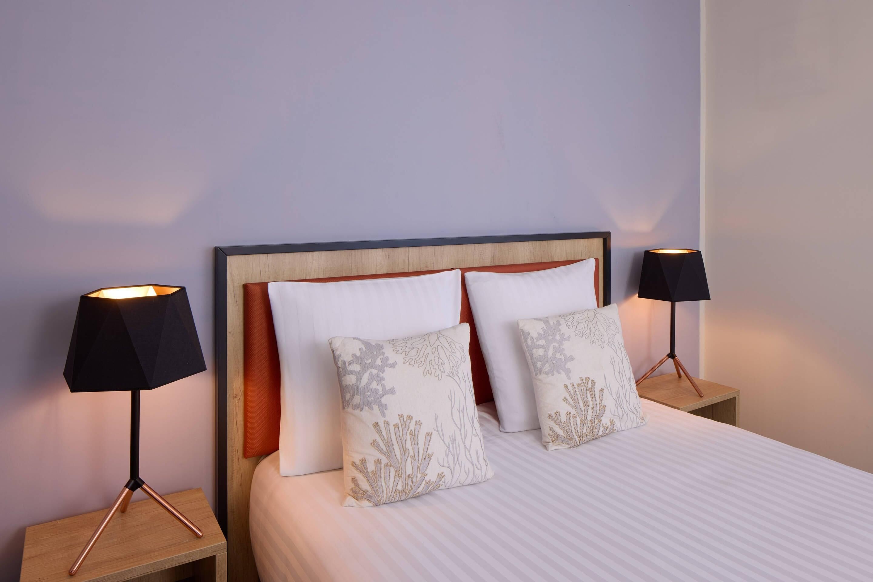 A neatly made bed with white bedding and two decorative pillows, flanked by two bedside tables with lamps, set against a plain wall.