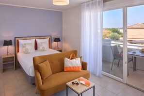 A hotel room with a bed, a sofa, a table with a fan and sunglasses, a nightstand with lamps, and a balcony with a table and chairs. - Quality Aparthotel Seyssinet (Seyssinet-Pariset)