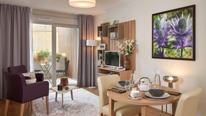 A modern living room with a dining area, a flat-screen TV, a bookshelf, and a large floral artwork on the wall. - Quality Aparthotel Seyssinet (Seyssinet-Pariset)