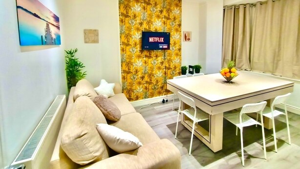 Een moderne woonkamer met een bank, een eettafel met stoelen, een aan de muur bevestigde televisie en tropische behang.