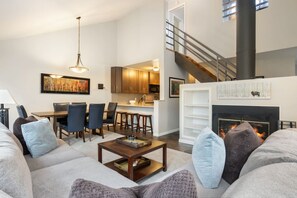 Ein modernes Wohnzimmer mit Sofa, Couchtisch und Kamin. Im Hintergrund befindet sich ein Essbereich mit Stühlen und eine Küche.