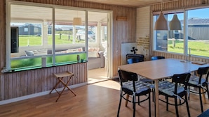 A room with a wooden floor, a dining table with chairs, a sofa, and a window with a view of a house and a car. - 3 bedroom beautiful home in Løkken (Løkken)