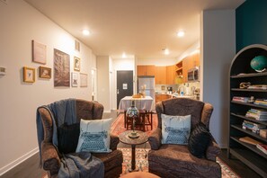 Uma sala de estar acolhedora com duas poltronas estampadas, uma mesinha redonda e uma estante repleta de livros.