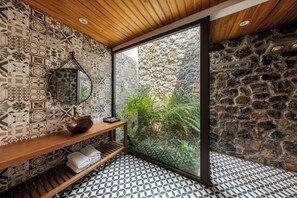 Een badkamer met patroontegels, een houten plank met handdoeken, een stenen muur en een groot raam met uitzicht op groen.